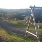 Spielplatz mit Schaukel und Klettergerüst auf grünem Gelände mit Wald im Hintergrund.