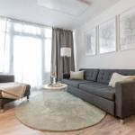 Wohnzimmer mit grauem Sofa, runden Teppich und Fenstern mit Vorhängen.