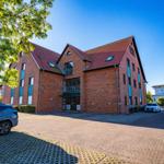 Rotes Backsteingebäude mit Dachterrassen und Parkplatz. Bäume und klare Himmel.