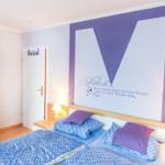 Schlafzimmer mit Bett, Nachttischen und Wanddekoration mit großem M und Text.