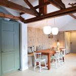 Dining area with wooden table, white chairs, and wood-paneled wall. Exposed beams and green cabinets.