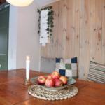 Wooden table with apple bowl and candle against wooden wall with cushions.
