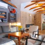 Living room with sofa, chairs, and wooden table. Wooden walls and ceiling beams. View into kitchen and bedroom.