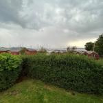 Green lawn area with hedge, behind it red wooden houses and sailboats on water.