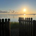 Sunset over calm water with sailboats and a fence in the foreground.