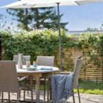 Terrasse mit Tisch, Stühlen und Sonnenschirm. Hintergrund: grüne Hecke und Holzzaun.