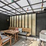 Gartenbereich mit Holztisch, Bank, Grill und Girlanden unter Dach.
