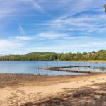 Seeufer mit Steg, Sandstrand und Wald im Hintergrund unter blauem Himmel.