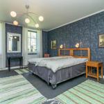 Schlafzimmer mit Bett, zwei Fenstern, Wandbildern und einem modernen Deckenlicht.