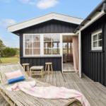 Wooden terrace with lounge chair, table and stools in front of black wooden house with windows.