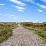 Gepflasterter Weg durch sandige Dünen unter blauem Himmel mit weißen Wolken.