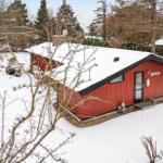 Red wooden house with snow-covered roof and garden area in winter.