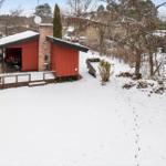 Red wooden shed with snow-covered roof and terrace in snowy garden.