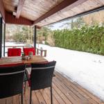 Deck with wooden table and chairs, snow in garden, green hedge in background.