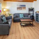Living room with two sofas, table, and wooden floor. Wall picture and plants visible.