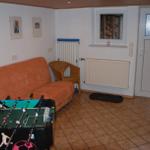 Raum mit orangefarbenem Sofa, Tischfußball und Wickerstuhl. Fenster und Tür sind sichtbar.