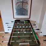 Fussballtisch mit Spielern und Tor. Wandbild darüber. Fliesenboden.