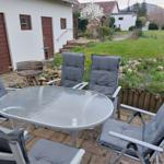 Terrasse mit Tisch, Stühlen und Grill. Hintergrund: Garten und Haus.