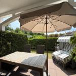 Terrasse mit Tisch, Stühlen und Sonnenschirm. Hintergrund: grüner Heckenzaun.