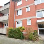 Rotes Backsteingebäude mit Balkon und Eingang. Grünflächen und Hecken vor dem Haus.