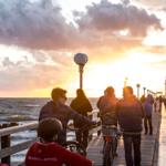 Besucher genießen Sonnenuntergang auf Steg mit Fahrrädern und Blick auf Meer.