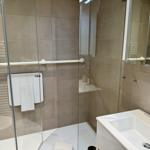 Duschkabine mit Handlauf und Sitzbank, daneben Waschbecken mit Spiegel.