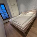Schlafzimmer mit Doppelbett, Schrank und Fenster. Holzboden und gemusterte Tapete.