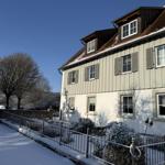 Wohnhaus mit Schneebedecktem Dach und Fenstern, umgeben von schneebedeckten Sträuchern und einem Zaun.