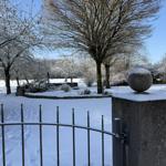 Schneebedeckter Garten mit Zaun und Steinbollard. Bäume und Sträucher sind mit Schnee bedeckt.