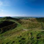 Green hills with a hiking trail and view of the sea under a blue sky.