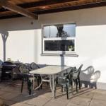 Terrace with table, chairs, and grill under wooden cover. Window with plant on sill.
