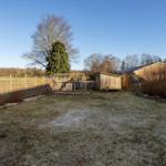 Backyard with frost-covered grass, wooden shed, and garden fence. Trees and shrubs surround the area.