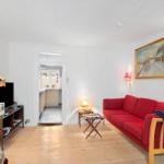 A living room with a red sofa, wooden floor, and view into the kitchen.