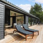 Deck with lounge chairs, table and chairs. Wood siding and glass facade. Background: forest.