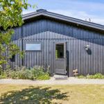 Black wooden house with entrance door and small window. Garden area with flowers and trees.