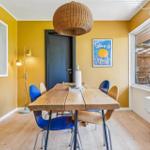 Dining room with wooden table, blue chairs, and yellow walls. Large windows and pendant light.