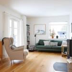 Living room with green sofa, wooden floor, and windows. A chair, bookshelf, and wood stove are visible.