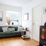Living room with green sofa, wooden floor, and wood stove. Window, pictures, and plants.