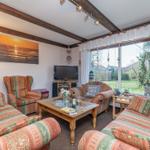 Wohnzimmer mit Sofa, Sessel und TV. Durchblick auf Garten durch große Fenster.