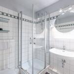 Badezimmer mit Dusche, Waschbecken und Spiegel. Weiße Fliesen und Glaswand.