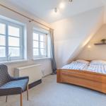 Doppelbettzimmer mit zwei Fenstern, Stuhl und Holzbett unter Dachschräge.