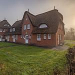 Rotes Backsteingebäude mit Strohdach und weißen Fenstern auf grünem Grund.