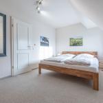 Schlafzimmer mit Dachschrägen, Holzbett, Spiegel und Fenster mit Vorhängen.