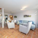 Wohn- und Essbereich mit Sofa, Sesseln und Holztreppe. Terrakottaboden und Wandbilder.