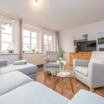 Wohnzimmer mit Sofa, zwei Sesseln, Glas-Tisch und Fernseher. Terrakotta-Boden und Fenster mit Vorhängen.