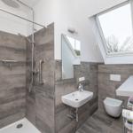 Badezimmer mit Dusche, Waschbecken und WC. Dachfenster und graue Fliesen.