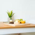 Holztisch mit Käse, Obst und Pflanze. Weiße Stühle und Wand im Hintergrund.