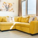 Gelber Ecksofa mit Kissen, Wandbild und Fenster mit Vorhängen.