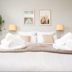 Das Bett mit weißer Bettwäsche, Kissen und aufgerollten Handtüchern. Zwei Bilder und eine Pflanze an der Wand.