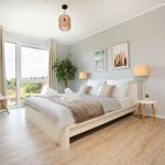 Schlafzimmer mit Bett, Nachttisch, Lampe und großem Fenster. Holzboden und Wandbilder.